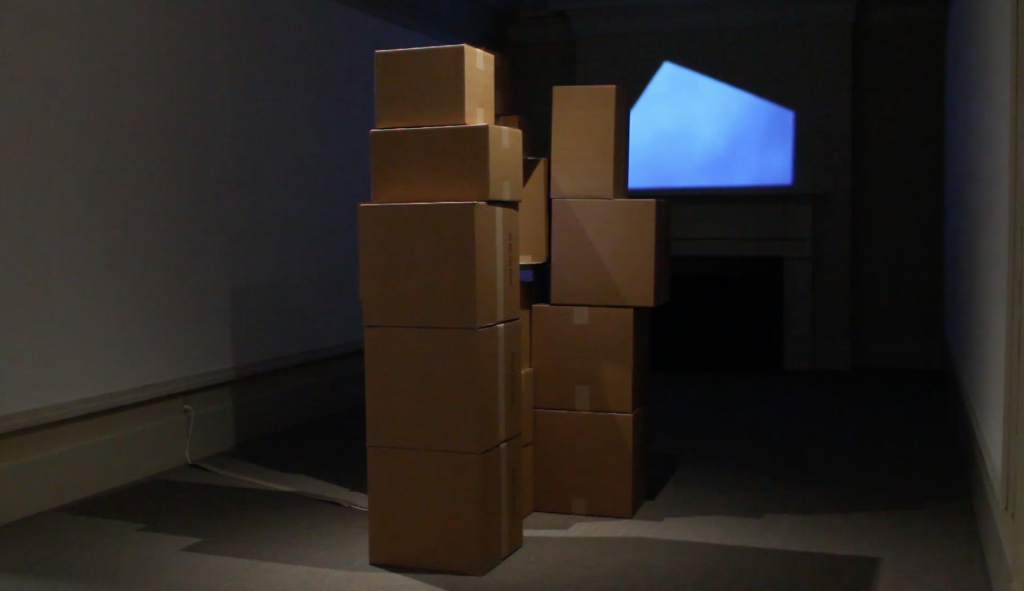 video still image of a stack of cardboard boxes in front of a video projection.